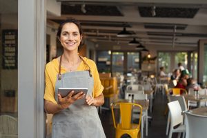 Small business owner in casual clothing and grey apron standing at entrance of coffee shop and looking at camera.